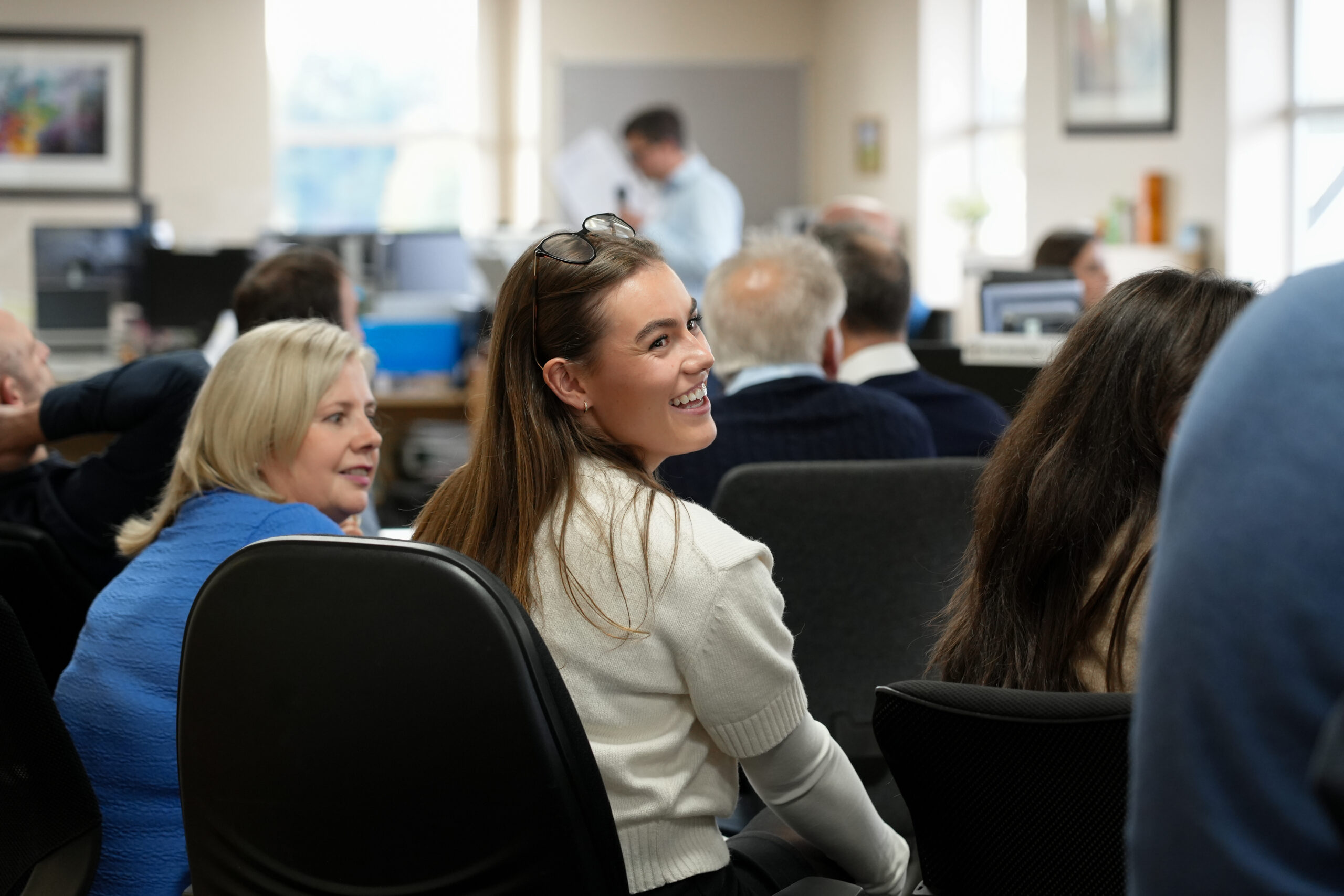 Group of colleagues in an office meeting room, seated and facing forward with one woman smiling, others visible in background