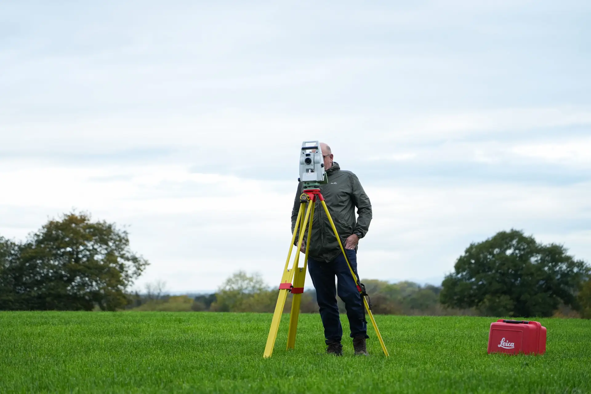 Surveyor using theodolite on tripod in open green field with trees in background and red equipment case nearby